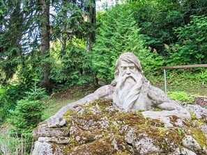 Eine steinerne Statue eines bärtigen Mannes auf einem moosbewachsenen Felsen in bewaldeter Landschaft.