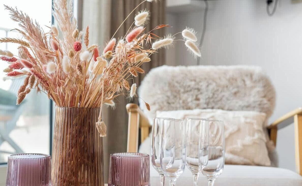 A room with a large window, a plush chair, a vase with dried plants, and a table with glasses.