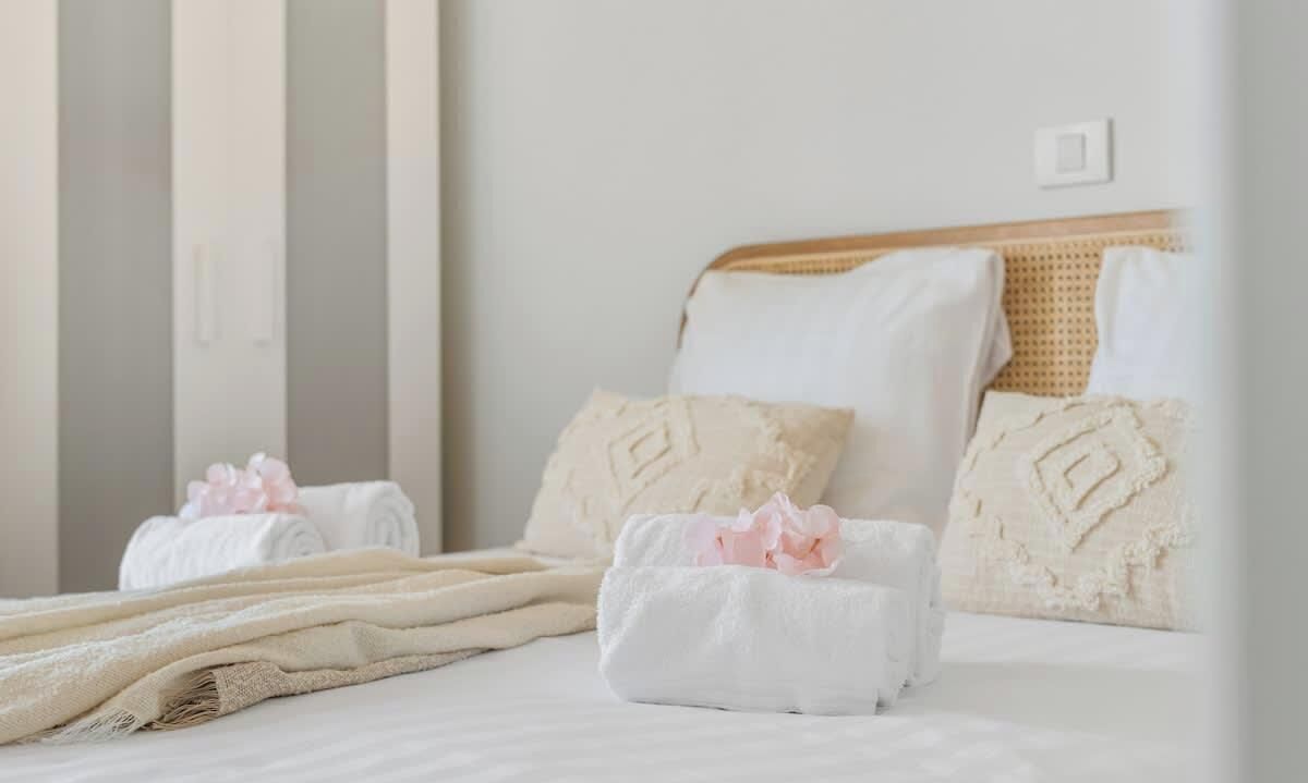 A neatly made bed with white linens and decorative pillows, a woven headboard, and a wall-mounted light switch.