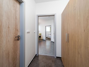 Ein Flur mit einer Holztür, einer weißen Wand und Blick in einen Raum mit Bett und Sessel.