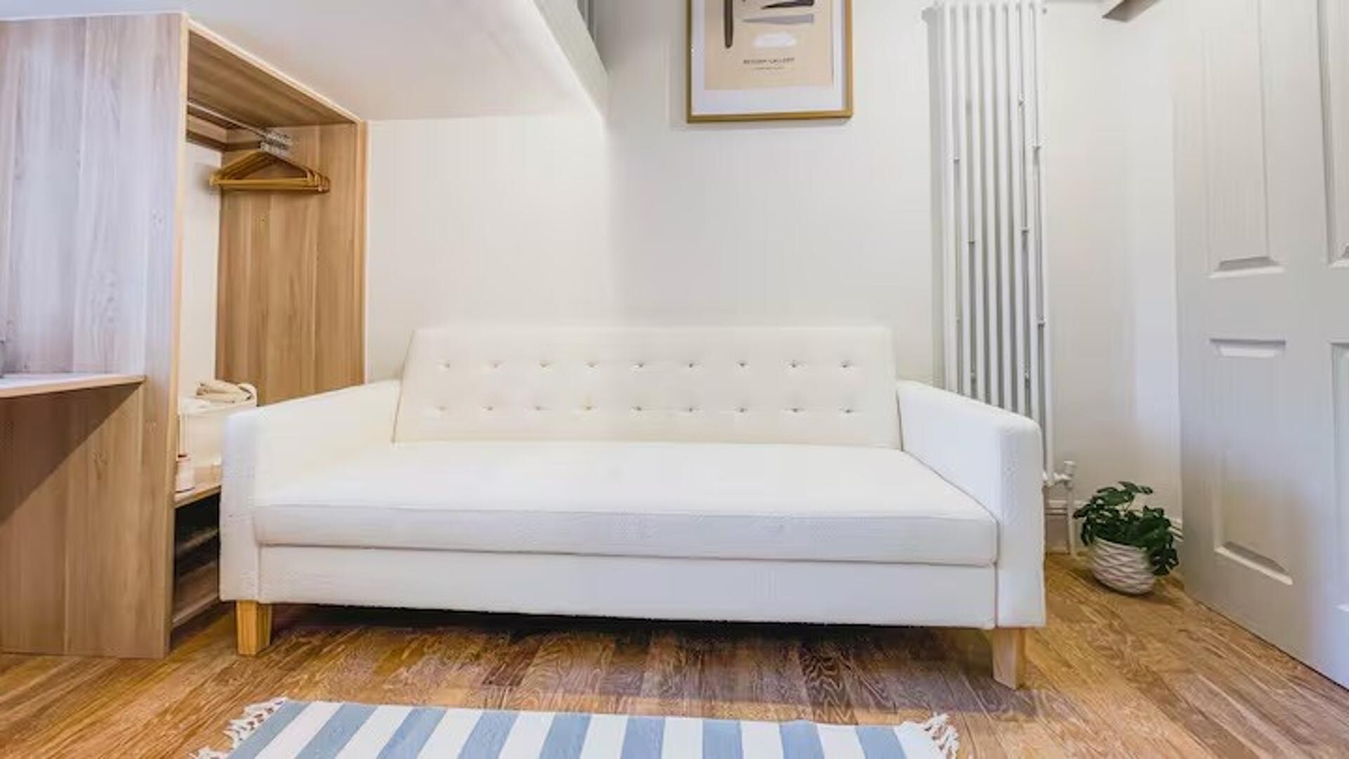 Un sofá blanco en una habitación con suelo de madera, un radiador y un cuadro enmarcado en la pared.