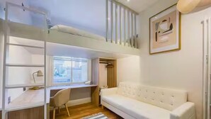 Ein Zimmer mit einem Etagenbett, einem Schreibtisch, einem Stuhl, einem weißen Sofa und einem Fenster.