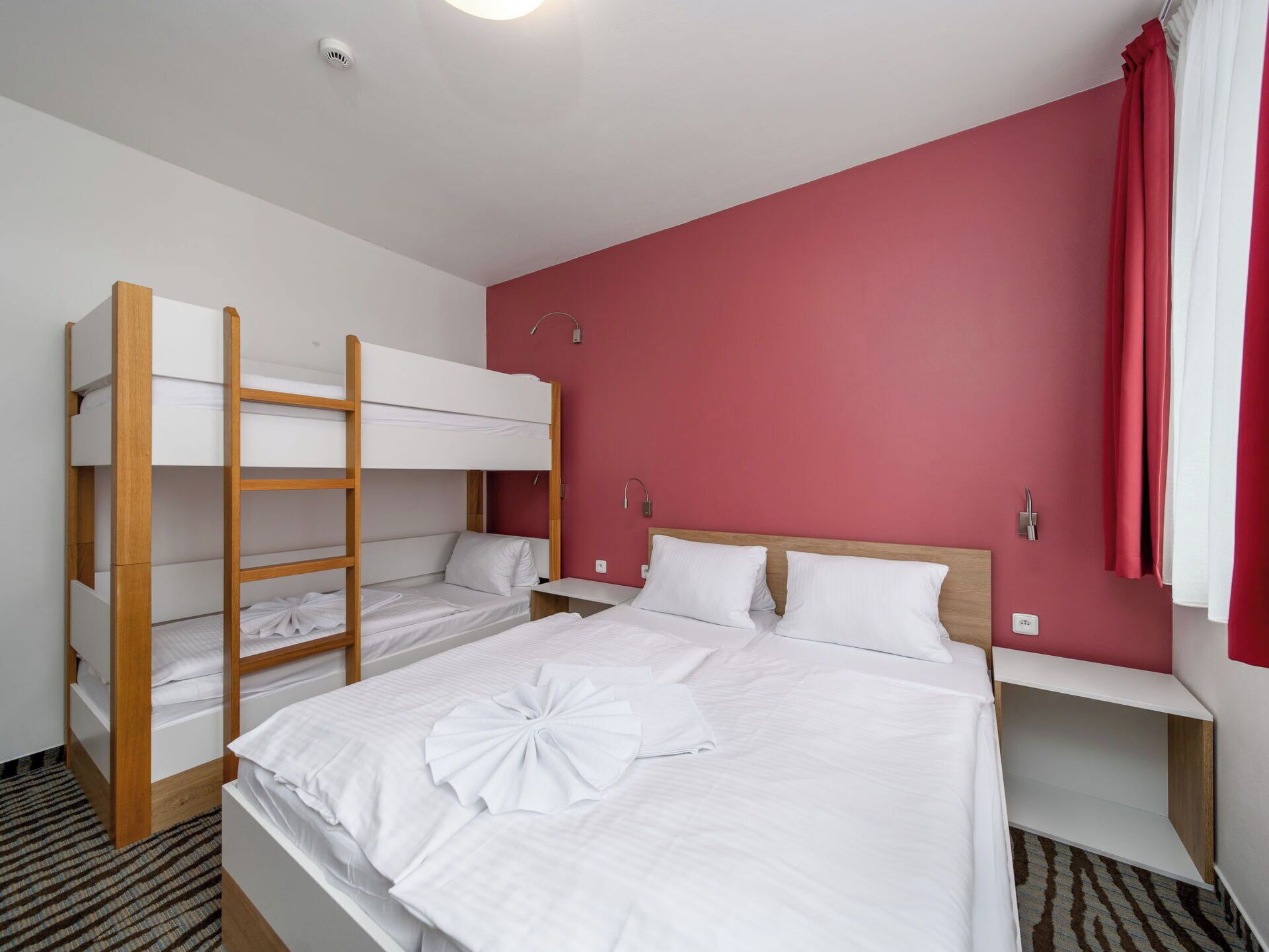 A hotel room with bunk beds, a large bed, and a red accent wall.