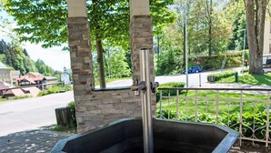 Eine öffentliche Wasserfontäne mit einem Metallauslauf und einem steinernen Sockel, umgeben von einem Metallzaun und Grünpflanzen.