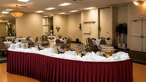 A banquet hall with a long table covered in white tablecloths, surrounded by chairs.