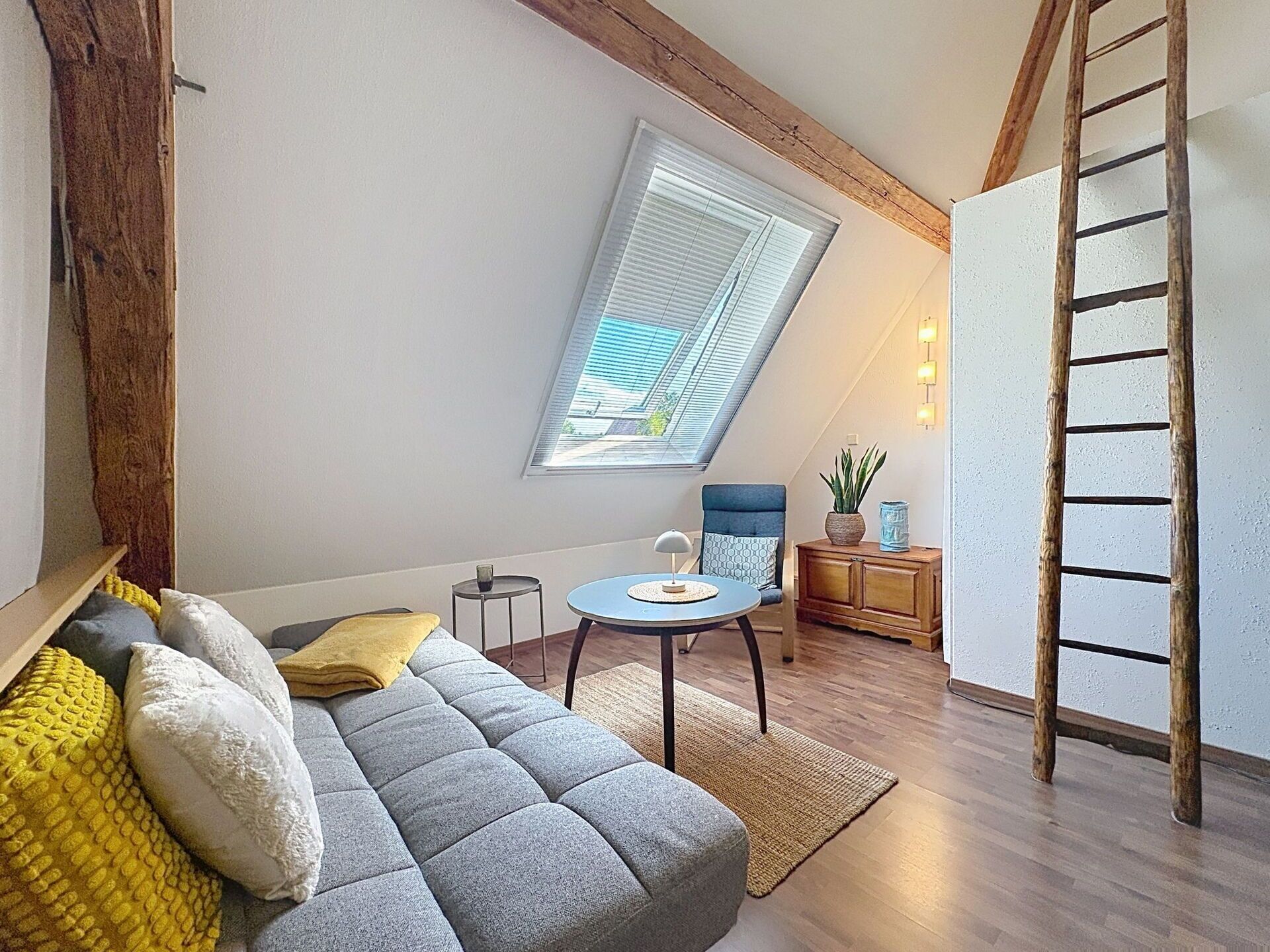 Un dormitorio tipo loft con techo inclinado, banco empotrado, una mesa redonda, una escalera y un ventanal con persianas.