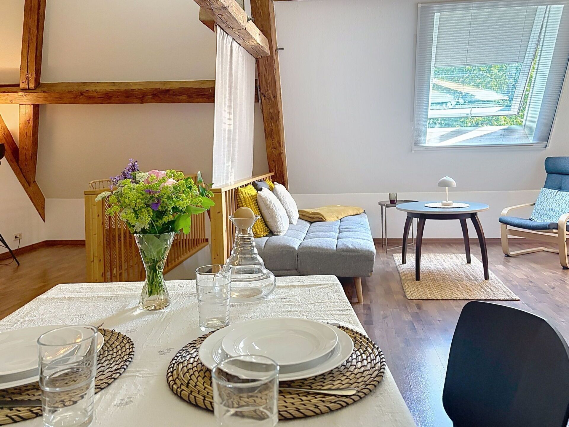 Un coin repas avec une table dressée pour quatre personnes, un canapé, une petite table avec une lampe et une fenêtre avec des stores.