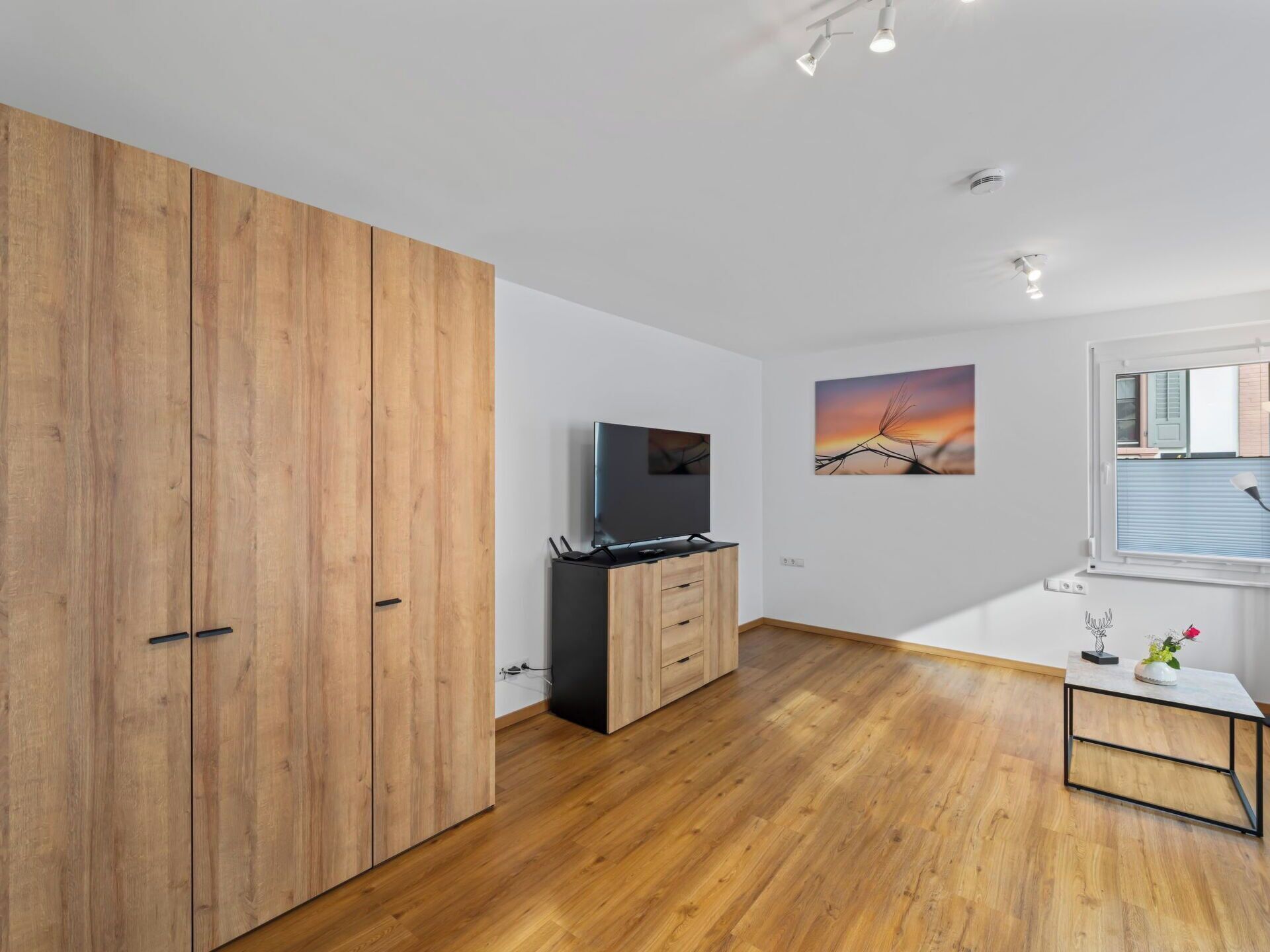 Ein modernes Zimmer mit Holzboden, einem Fernseher, einem Holzschrank, einem kleinen Tisch mit einer Vase und einem Fenster mit Jalousien.