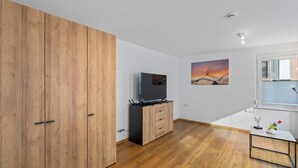 Ein modernes Zimmer mit Holzboden, einem Fernseher, einem Holzschrank, einem kleinen Tisch mit einer Vase und einem Fenster mit Jalousien.
