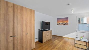 Ein modernes Zimmer mit Holzboden, einem Fernseher, einem Holzschrank, einem kleinen Tisch mit einer Vase und einem Fenster mit Jalousien.