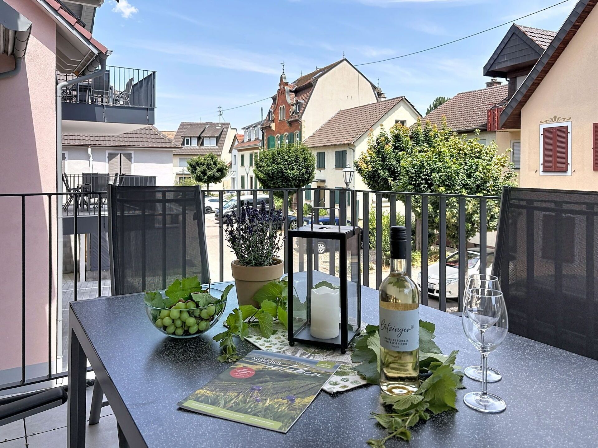 Een balkon met een eettafel, met een schaal druiven, een kaars, een fles wijn en een glas.