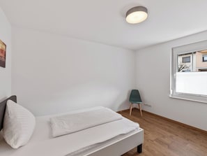 Ein schlicht eingerichtetes Schlafzimmer mit Bett, Stuhl, Fenster und Deckenleuchte.