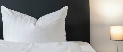 A neatly made bed with white linens and pillows, a dark headboard, and a bedside table with a lamp.