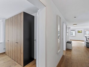 Ein modernes Interieur mit einem Holzschrank, einer weißen Tür und einem Flur, der in einen Raum mit einem grauen Sofa und einem kleinen Tisch führt.