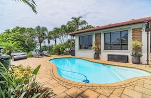A swimming pool with a curved edge, a patio area with lounge chairs, and a house with large windows and a tiled roof.