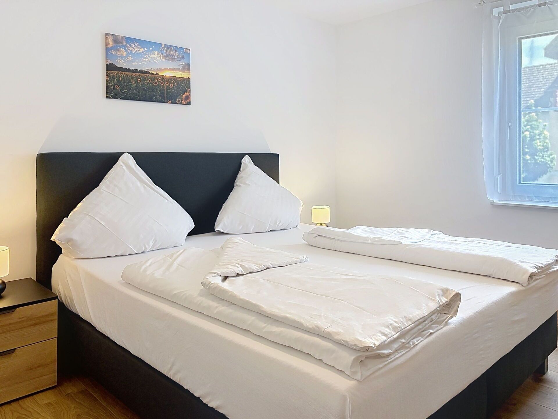 A neatly made bed with white linens and pillows, a bedside table with a lamp, and a window with a view of greenery.