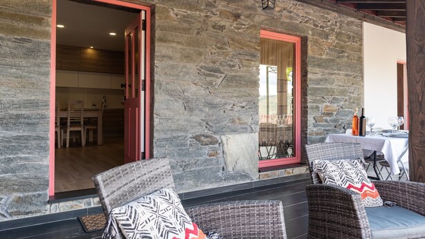 A stone exterior wall with a red-framed window and a door, wicker chairs with patterned cushions, and a table with a bottle and glasses.