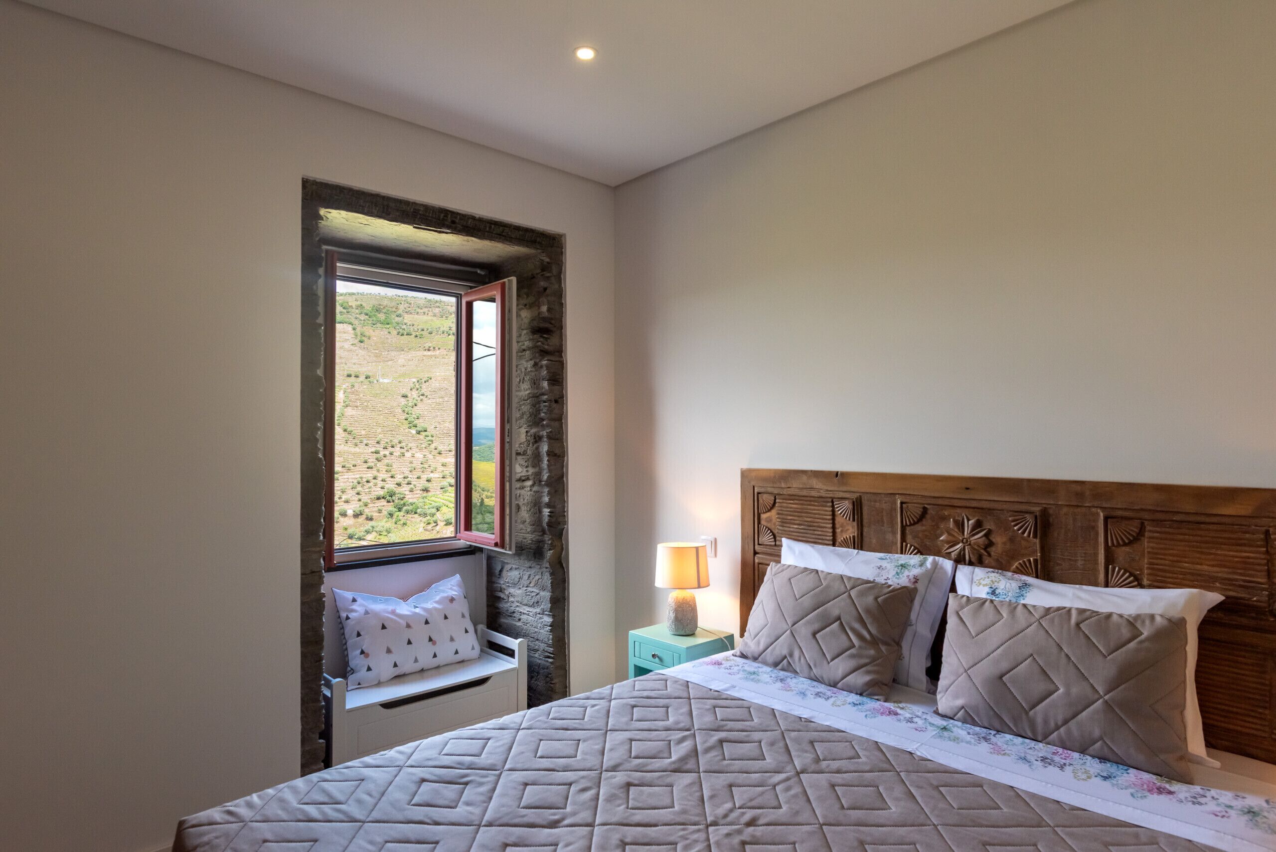 Un dormitorio con cama, mesita de noche, lámpara, ventana con vistas y cabecera de madera.