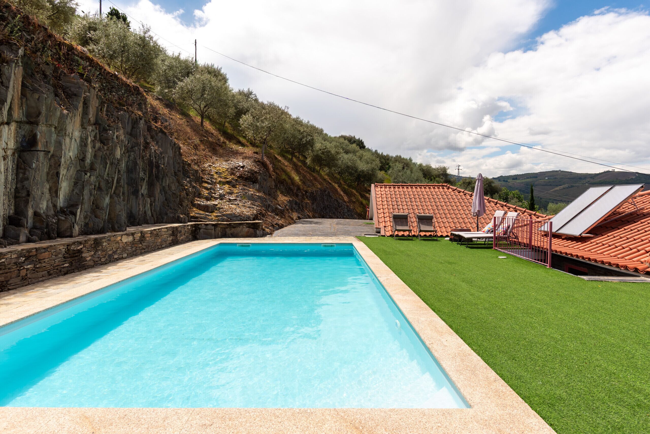 Una piscina rectangular con una pared de piedra en un lado y una zona verde con un edificio de techo rojo al fondo.