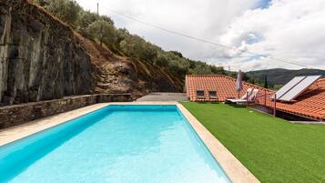 Una piscina rectangular con una pared de piedra en un lado y una zona verde con un edificio de techo rojo al fondo.