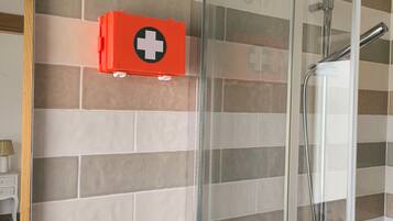 Un espace douche délimité par une paroi en verre, une trousse de premiers secours rouge fixée au mur, et un mur recouvert de carreaux formant un motif.