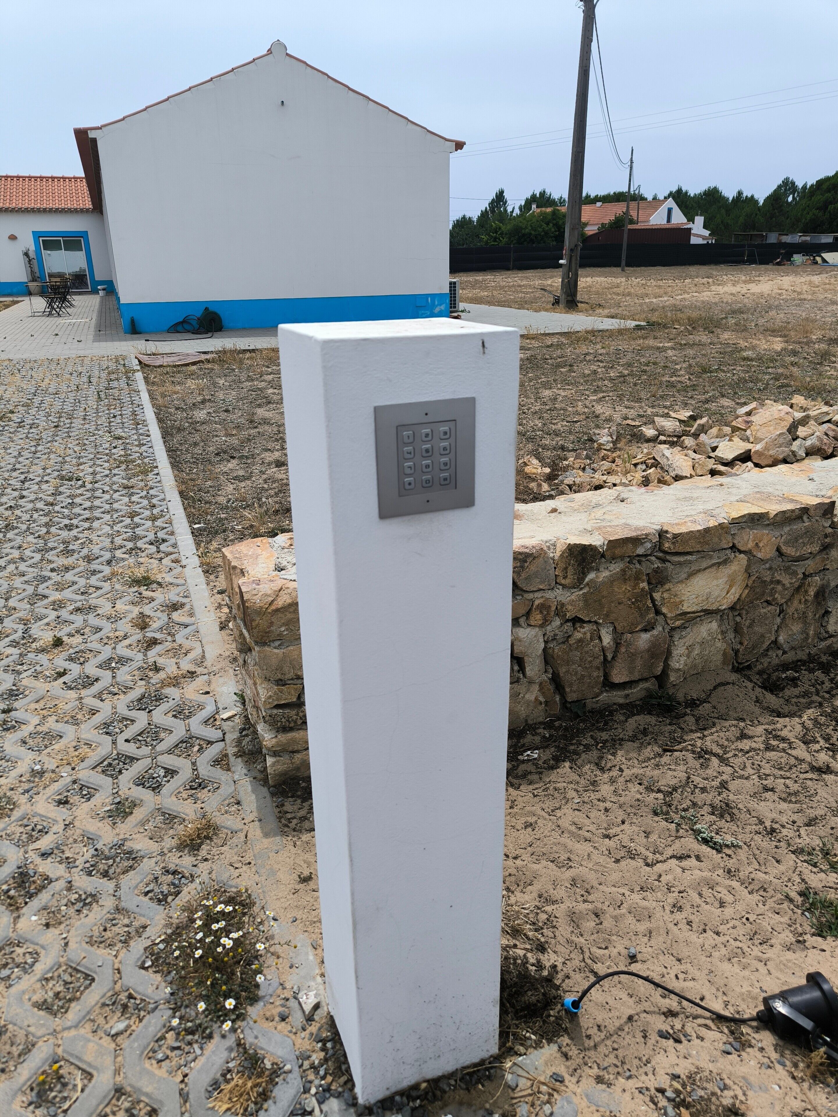 A white electrical box with a keypad, a stone wall, and a paved area with a building in the background.