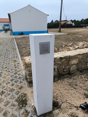 A white electrical box with a keypad, a stone wall, and a paved area with a building in the background.