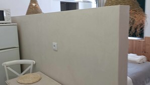 Ein Zimmer mit einem weißen Kühlschrank, einer Mikrowelle darüber, einem weißen Stuhl, einer gewebten Stuhlmatte und einem Bett mit weißer Decke.