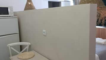 Una habitación con un refrigerador blanco, un microondas encima, una silla blanca, un tapete de silla tejido y una cama con una manta blanca.