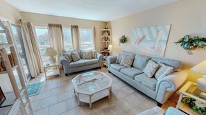 A living room with two grey sofas, a glass coffee table, a bookshelf, and a painting on the wall. - Coastal 1BR Condo at Emerald Isle | Steps to Beach, Pool, Tennis & Sleeps 4 (Fort Walton Beach)