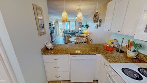A kitchen with white cabinets, a dishwasher, a stove, and a dining area with a table and chairs. - Coastal 1BR Condo at Emerald Isle | Steps to Beach, Pool, Tennis & Sleeps 4 (Fort Walton Beach)