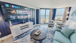 Ein modernes Hotelzimmer mit einem großen Fernseher, der verschiedene Streaming-Dienste anzeigt, einem Balkon mit Meerblick und einer gemütlichen Sitzecke mit einem Teppich im Marmor-Design.