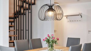 Uma sala de jantar com uma mesa de madeira, cadeiras cinzas, um vaso com flores e uma luminária pendente singular.