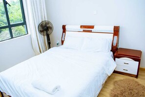 Ein ordentlich bezogenes Bett mit weißen Leinen, ein Nachttisch mit Schubladen, ein kleiner Holfnachtisch, ein an der Wand befestigter Ventilator und ein Fenster mit Vorhängen.