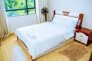 A single bed with white bedding, a bedside table with drawers, a fan, and a window with curtains. - Enjoyable 1-bedroom apartment near vibrant Nairobi city center (Nairobi)