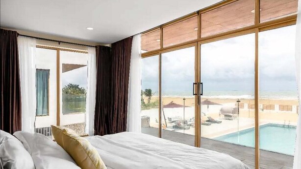 A bedroom with a large bed, a view of a pool and beach, and a sliding glass door.