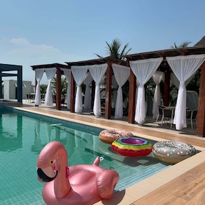 Una piscina amb flamencs i boies de forma de rosella inflables, envoltada d'zones d'estar amb seients coberts i cortines blanques.