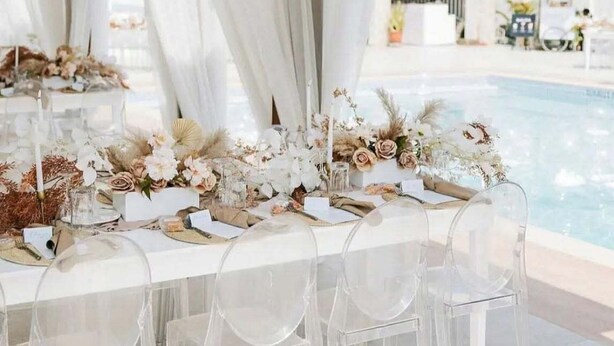 A poolside dining area with a table set for a meal, clear chairs, and a backdrop of white curtains and hanging lights.