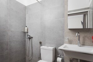 A bathroom with grey tiles, a white toilet, a shower with a handheld showerhead, a sink with a faucet, and a towel rack. - Koutelo by The Sea (Sfakia)