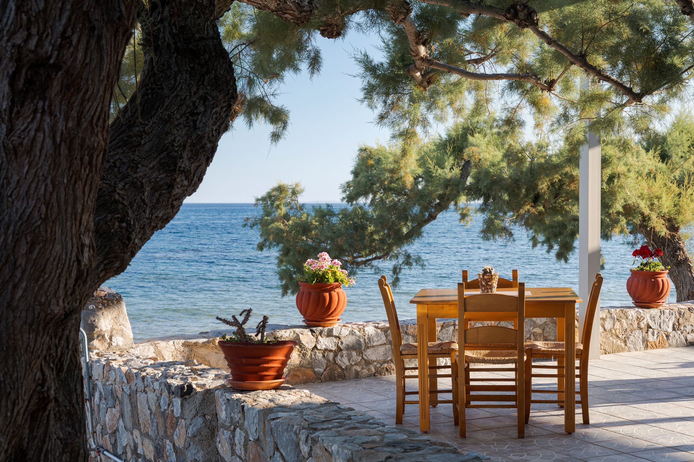 A stone wall, wooden table, and chairs by a body of water with potted plants.