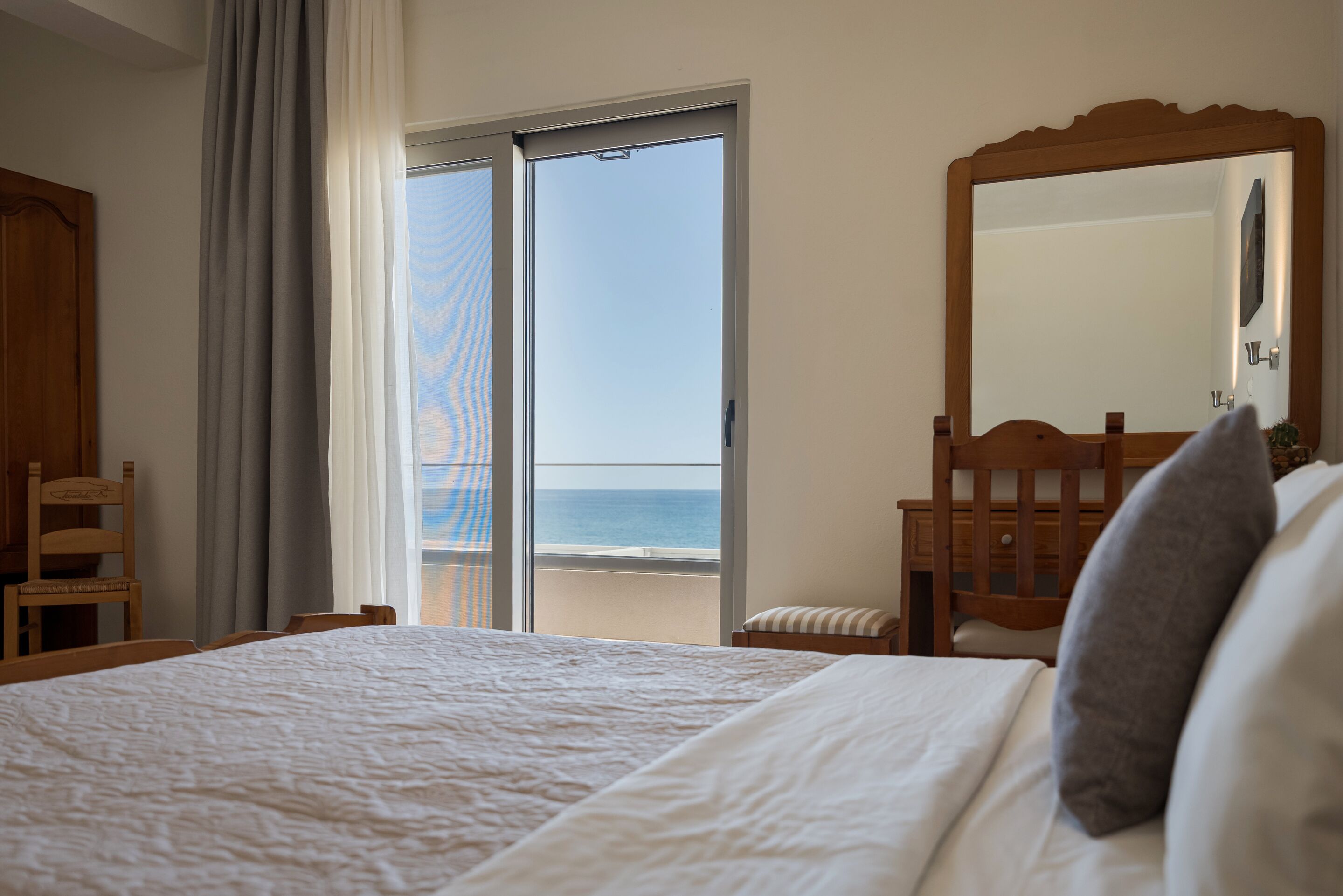 A hotel room with a bed, a chair, a small table, a mirror, and a view of the sea through a sliding glass door.