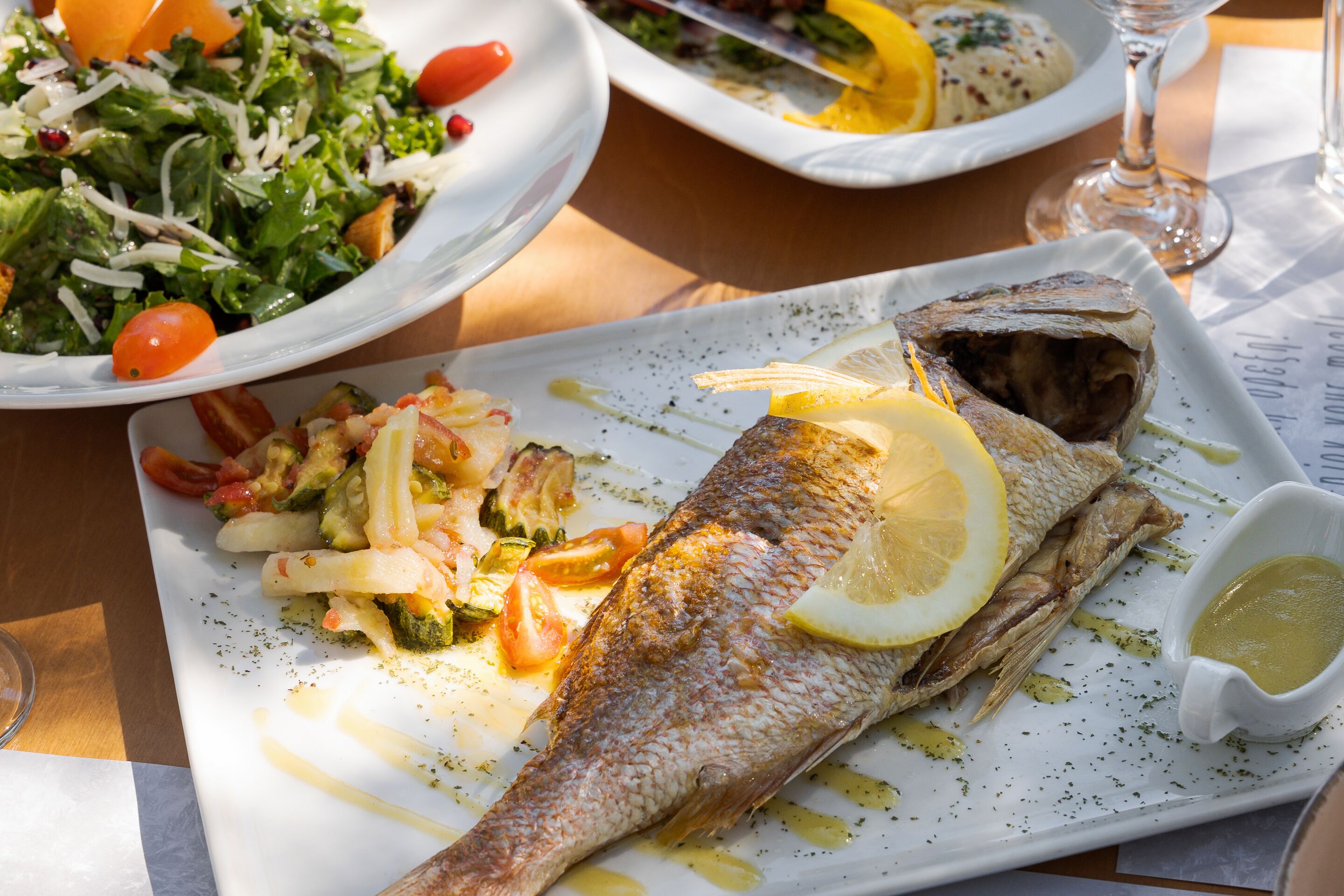 A dining table with a plate of fish, vegetables, and a small bowl of sauce.