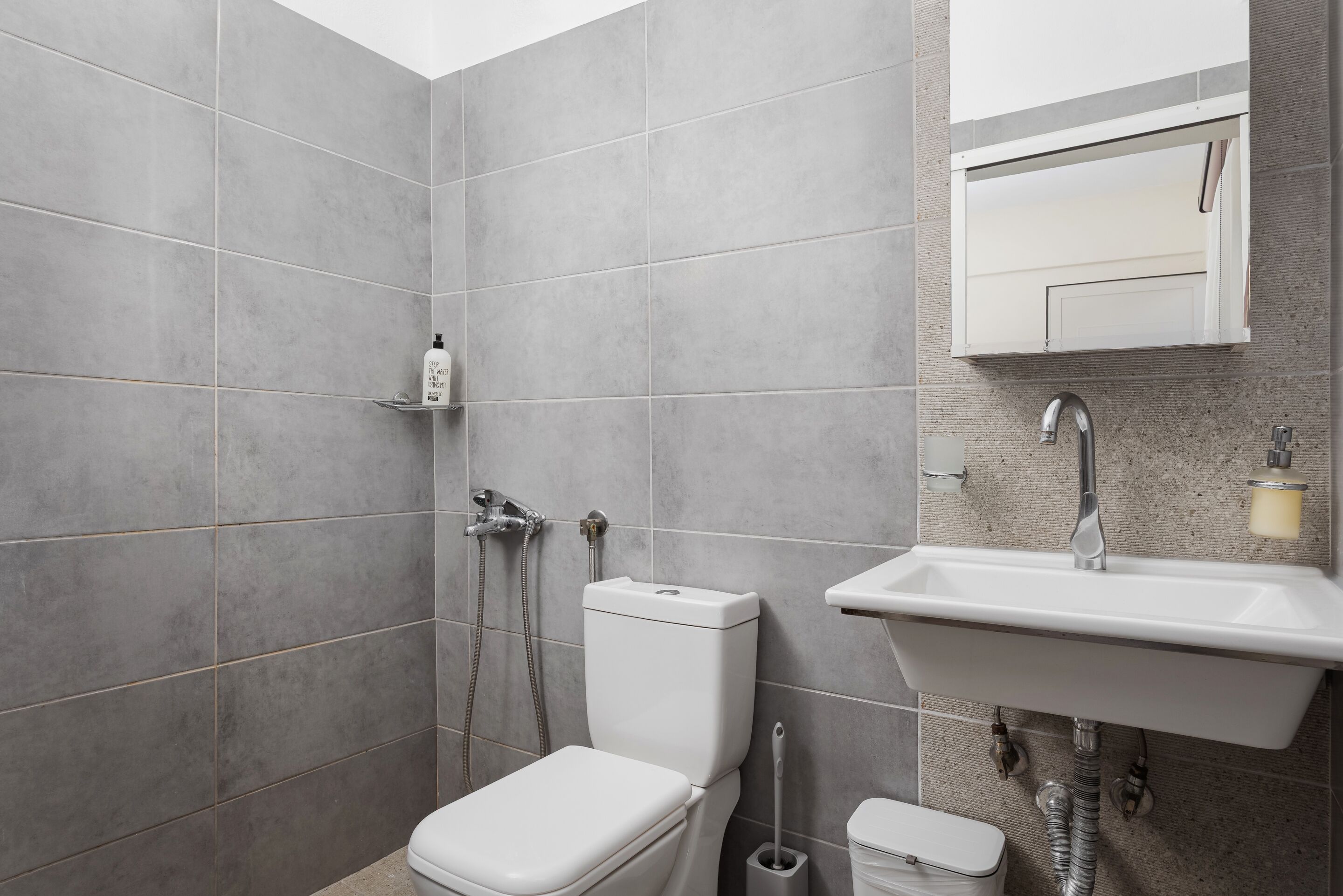 Ein modernes Badezimmer mit grauen Fliesen, einer weißen Toilette, einer Dusche mit Duschbrause, einem Waschbecken mit Spiegel darüber und einem Seifenspender an der Wand.