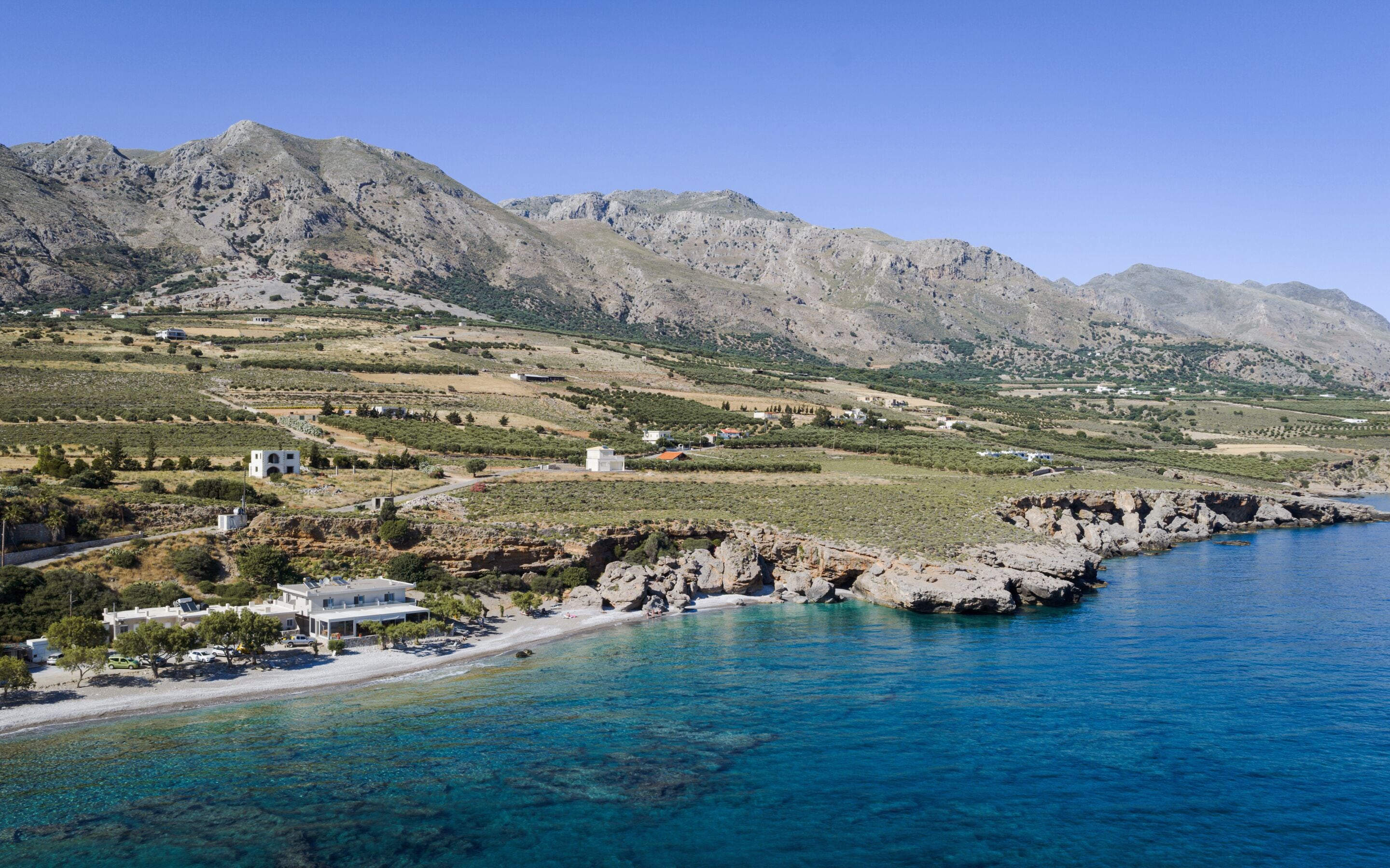 A coastal area with a beach, clear blue sea, rocky coastline, and mountainous background.