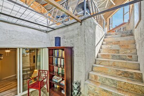 Una escala de pedra, una prestatgeria de fusta i una cadira vermella en una habitació amb paret de formigó i un finestral.