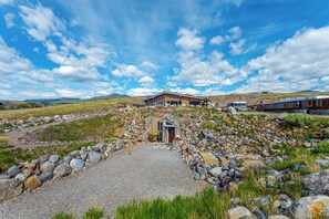 Una casa moderna con camino de grava, un pequeño patio y montañas al fondo.