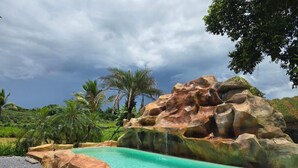 A swimming pool with a rocky waterfall feature, surrounded by palm trees and greenery. - My Paradise Private Resort Chiang Rai (Chiang Rai)