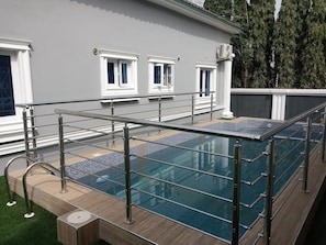 A swimming pool with glass panels, metal railings, and a tiled deck. - Romford 64 Apartment (Galadimawa)