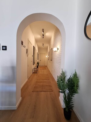 A hallway with wooden flooring, white walls, and an arched doorway. There are framed pictures on the walls, a potted plant, and a light fixture on the ceiling. - St. Julian's Apartment 3Bed - Near Beach&Paceville (Saint Julian's)