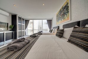 Quarto de hotel moderno com uma cama grande, televisão, varanda e vista para a cidade.